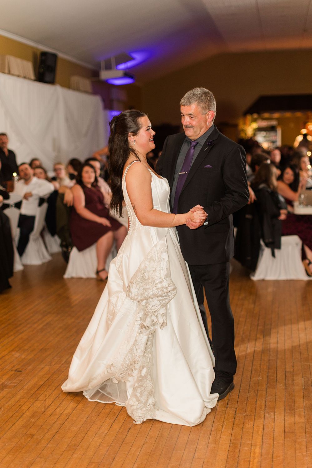 Smiling bride in a lace-trimmed white wedding dress dances with a gray-haired man in a dark suit as guests look on at the reception.