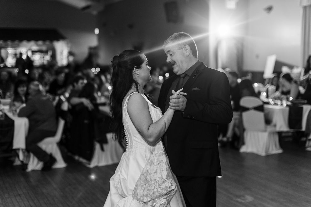 A bride in a white gown shares a first dance with a smiling man in a dark suit as wedding guests watch from tables in the background.