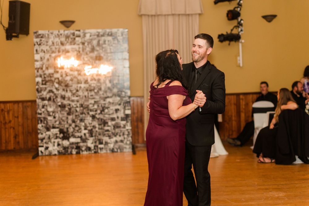 Man in a dark suit dances with a woman in a burgundy dress on a wooden floor at a reception with a photo wall backdrop.