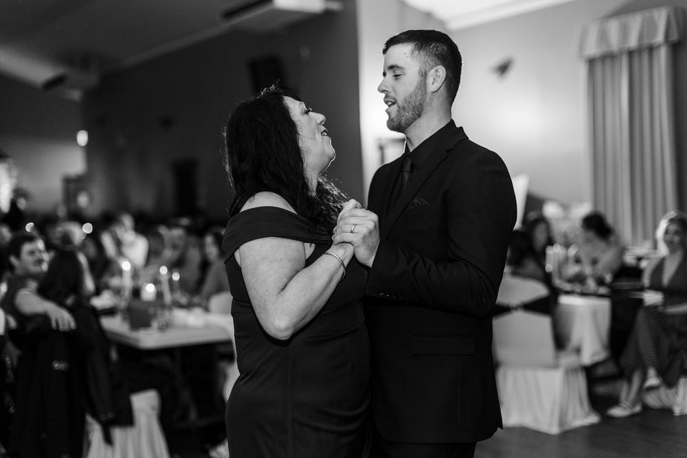 A woman in a black dress dances closely with a man in a dark suit and tie, holding hands, while wedding guests sit at tables behind them.