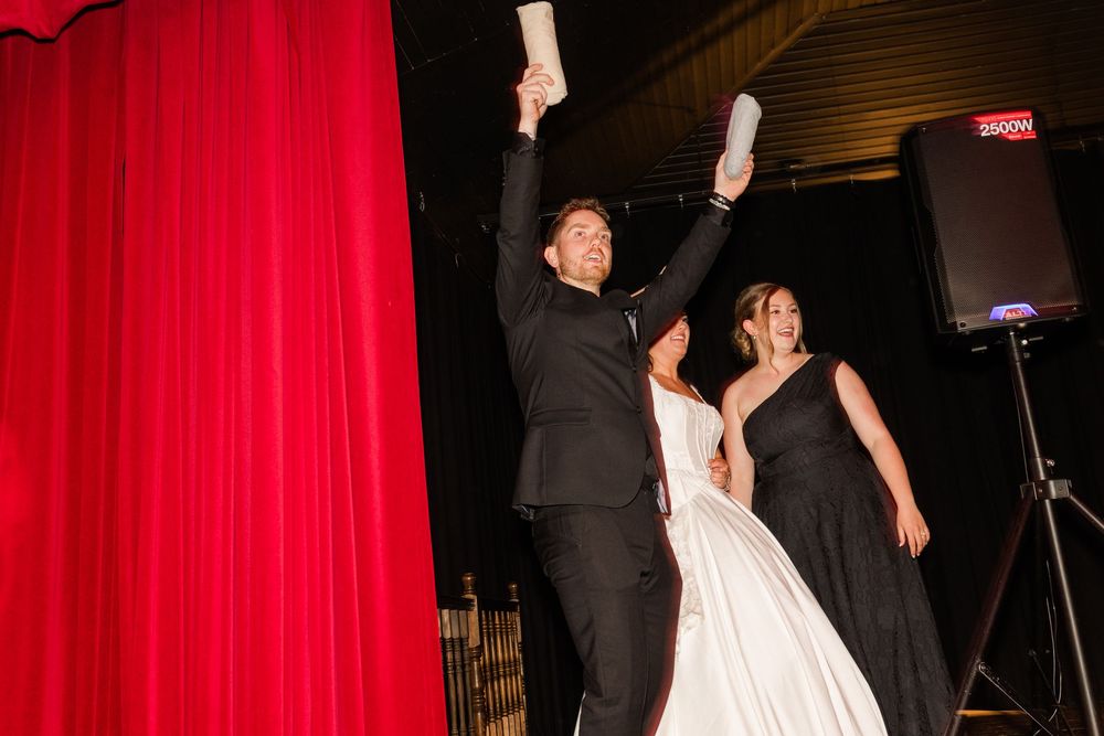 A groom in a black suit raises a paper in each hand beside a bride in a white gown and a woman in black near red curtains.