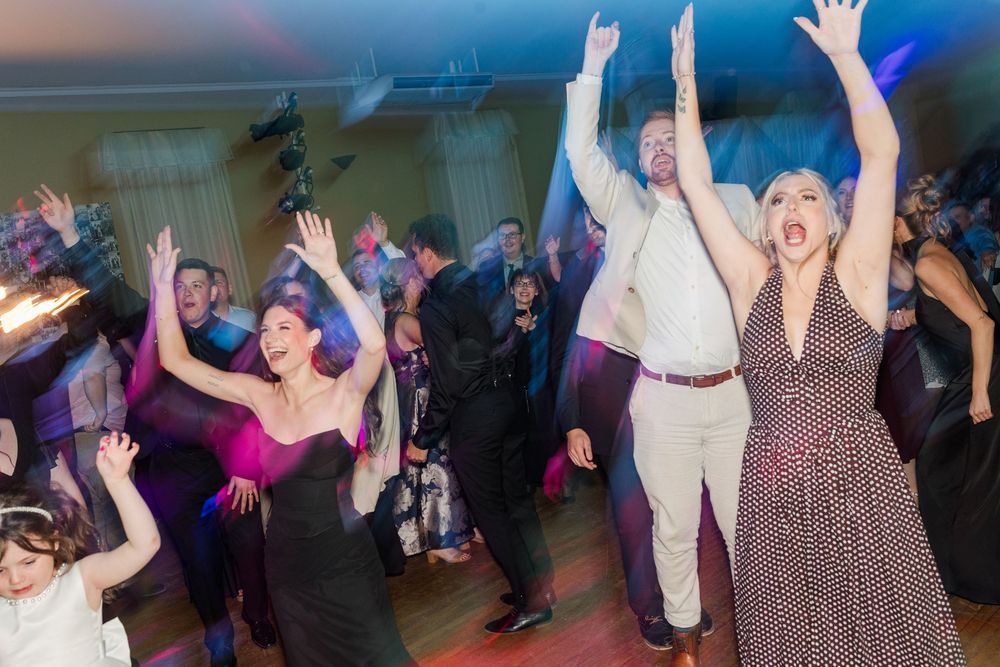 Guests dance joyfully with arms raised on a crowded wedding reception dance floor, with a woman in a polka dot dress in the foreground.