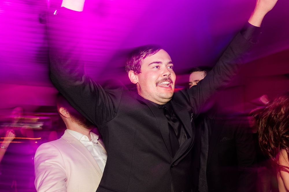 Smiling man in a black suit raises his arms and dances energetically under vibrant pink and purple lighting at a wedding reception.
