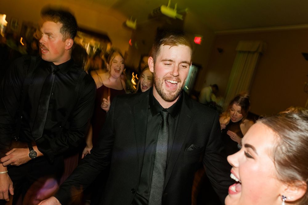 Smiling man in a dark suit and tie dances joyfully with laughing guests at a lively indoor wedding reception party.