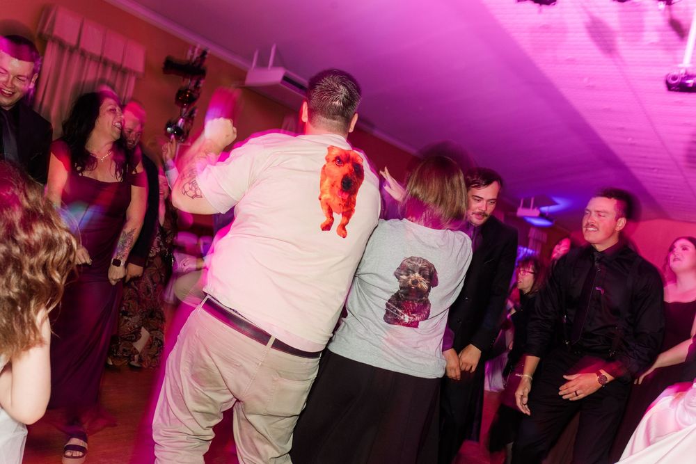 Guests dance at a lively indoor party, with two people in the foreground wearing casual shirts printed with dog graphics.