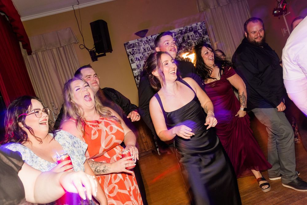A group of cheerful guests in colorful formal dresses and dark attire laugh and dance together at a lively indoor wedding reception.