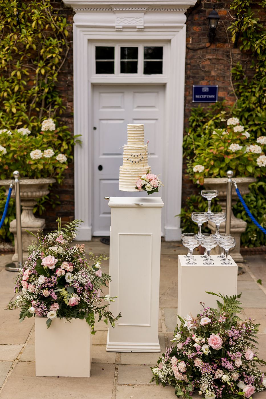 Elegant wedding cake display on white pedestal with champagne tower and pink floral arrangements outside a venue door.