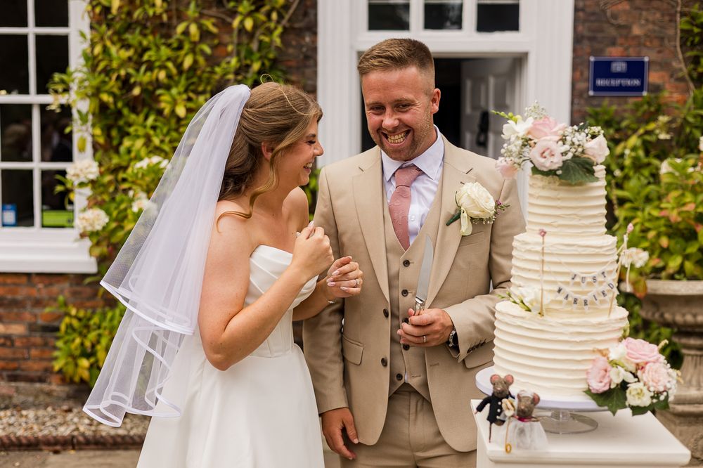 Bride and groom laughing together beside a white wedding cake outdoors at a reception venue.