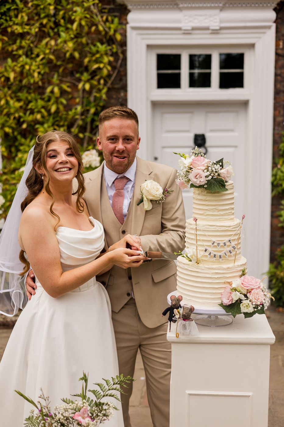 Bride and groom smiling while cutting a four-tier white wedding cake decorated with pink flowers outdoors.