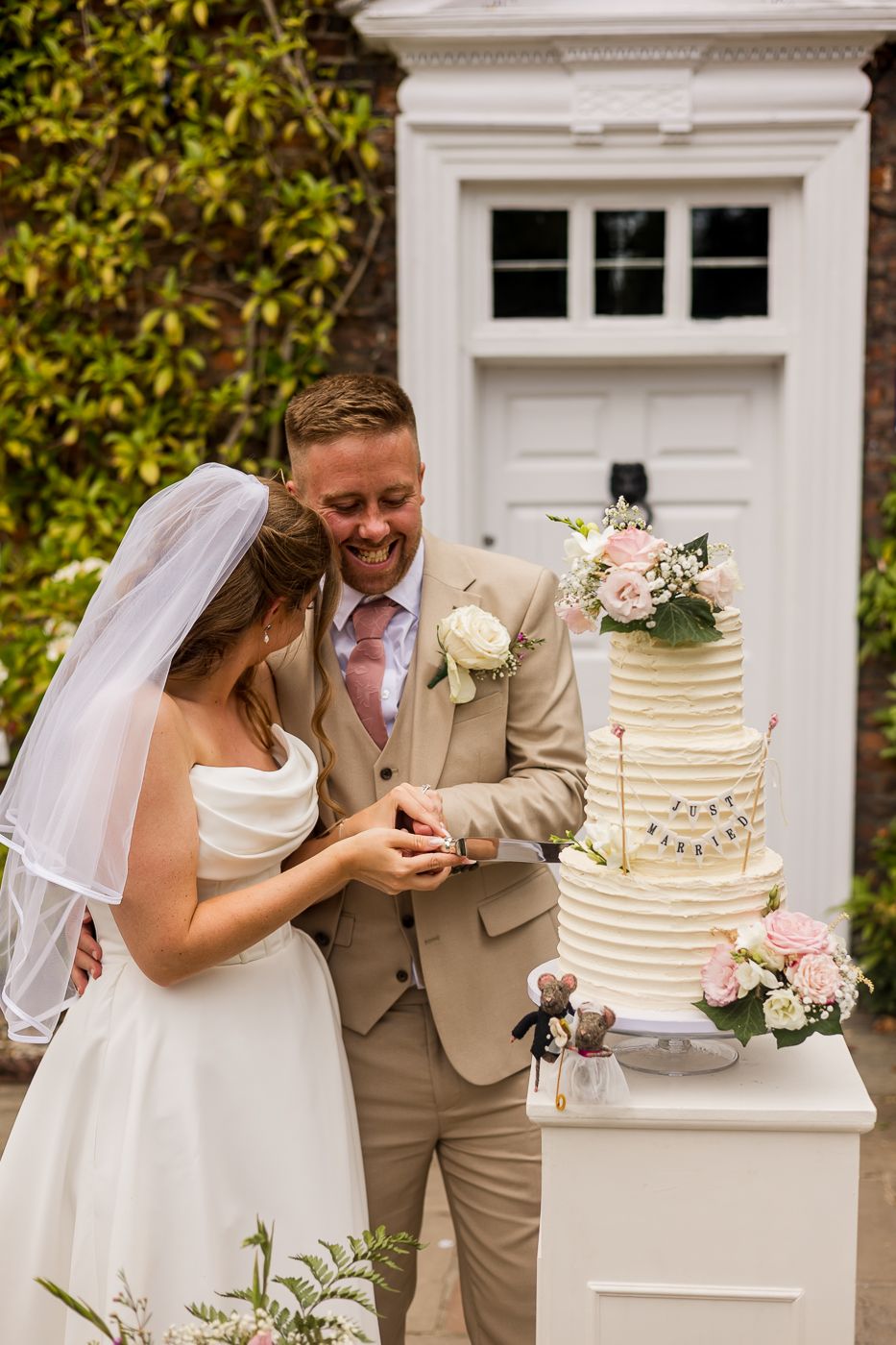 Bride and groom cutting their elegant tiered wedding cake adorned with pink roses outdoors on a sunny day.