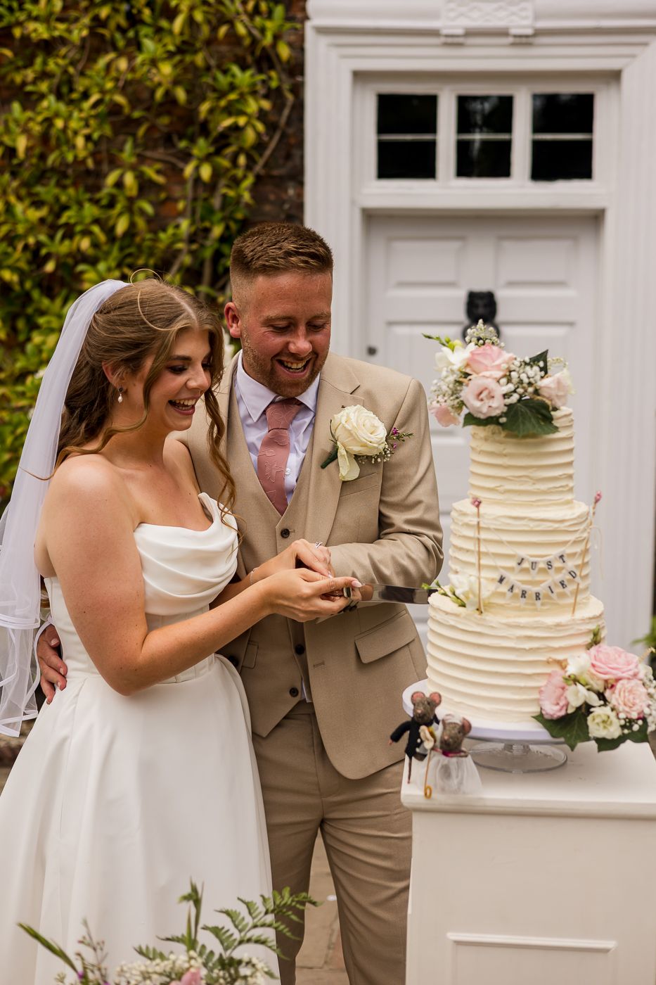 A smiling bride and groom in a beige suit cut their elegant tiered wedding cake outdoors near a white door.