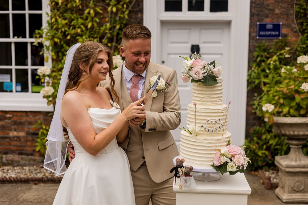 Bride and groom smiling while cutting a elegant white tiered wedding cake outdoors at a charming venue.