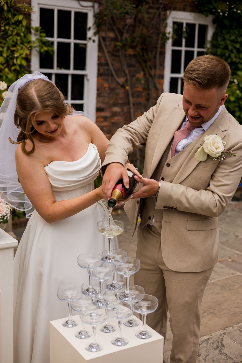 Bride and groom pouring champagne tower at wedding reception outdoors, wearing white gown and beige suit.