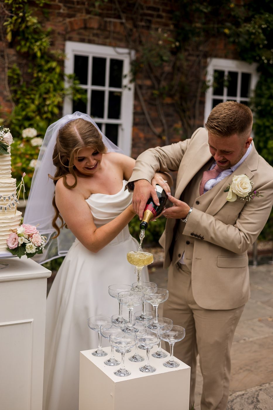 Bride and groom pouring champagne tower at outdoor wedding reception, wearing white gown and beige suit.