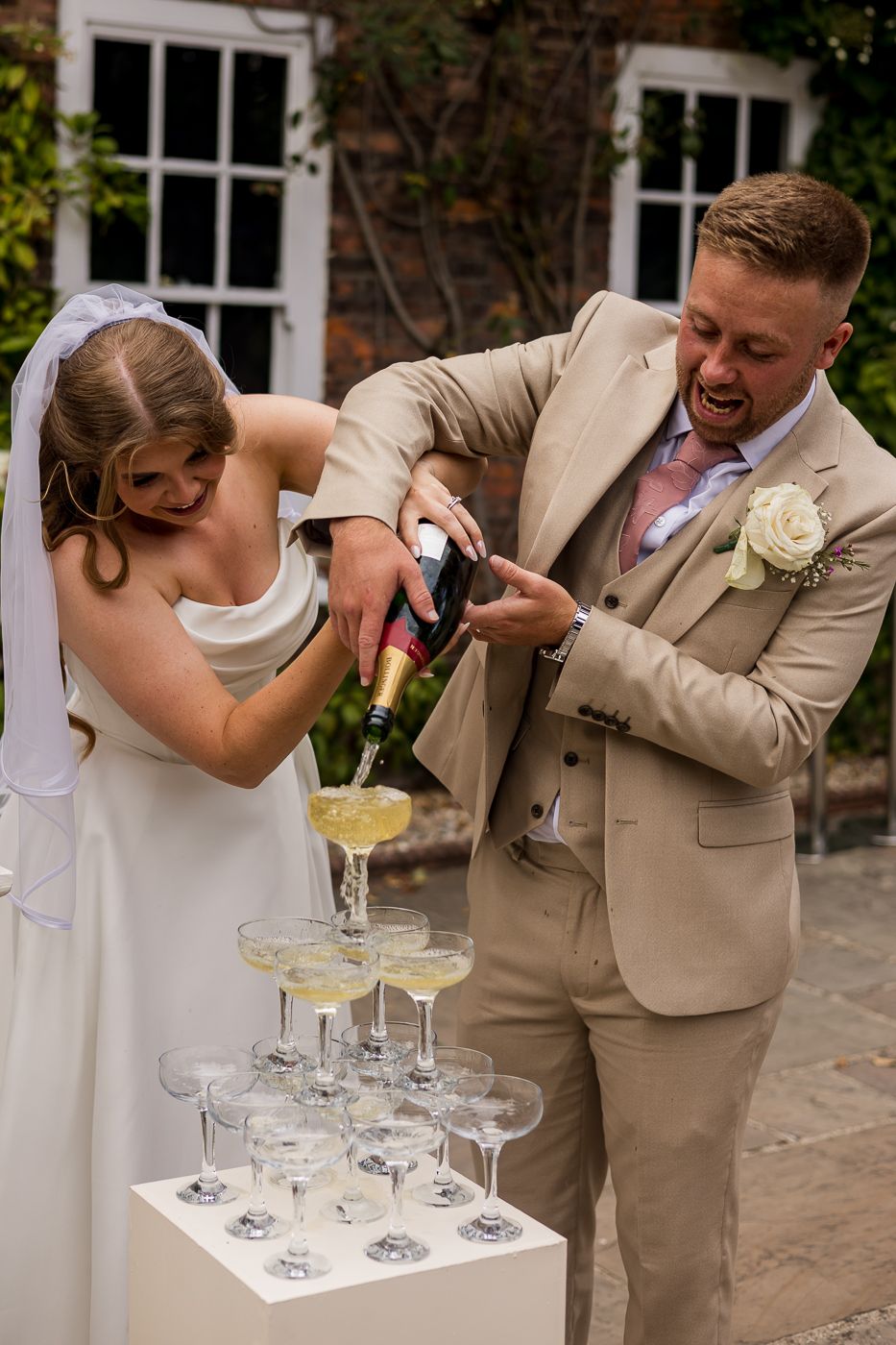 Bride and groom pouring champagne tower at wedding reception outdoors.