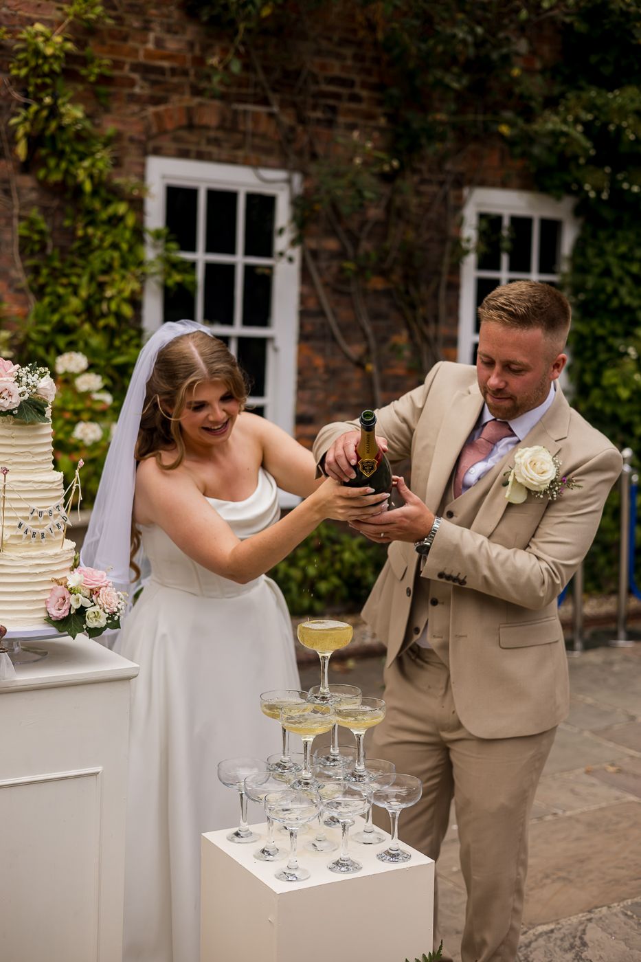 Bride and groom pouring champagne tower at outdoor wedding reception near brick building.
