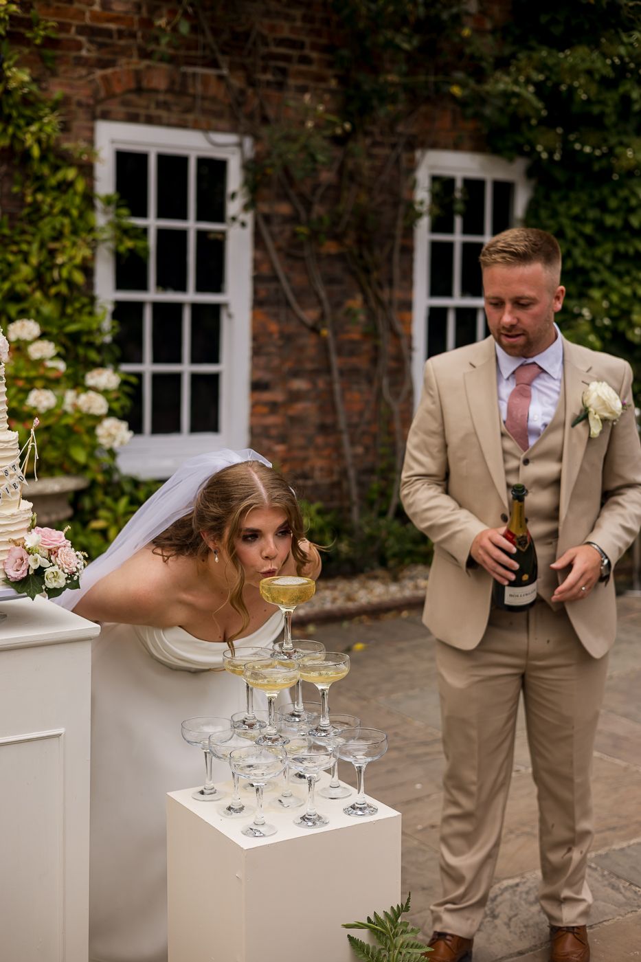 Bride and groom at champagne tower during outdoor wedding reception at brick building venue.