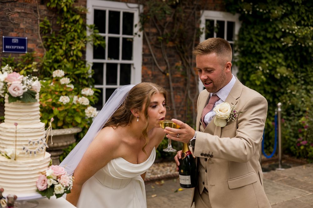 Bride and groom sharing champagne while cutting wedding cake outdoors at elegant venue.
