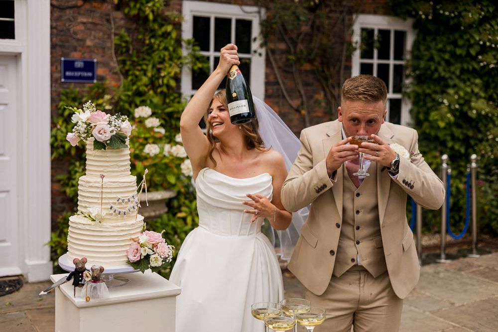 Bride holding champagne bottle aloft while groom drinks at outdoor wedding reception with tiered cake.