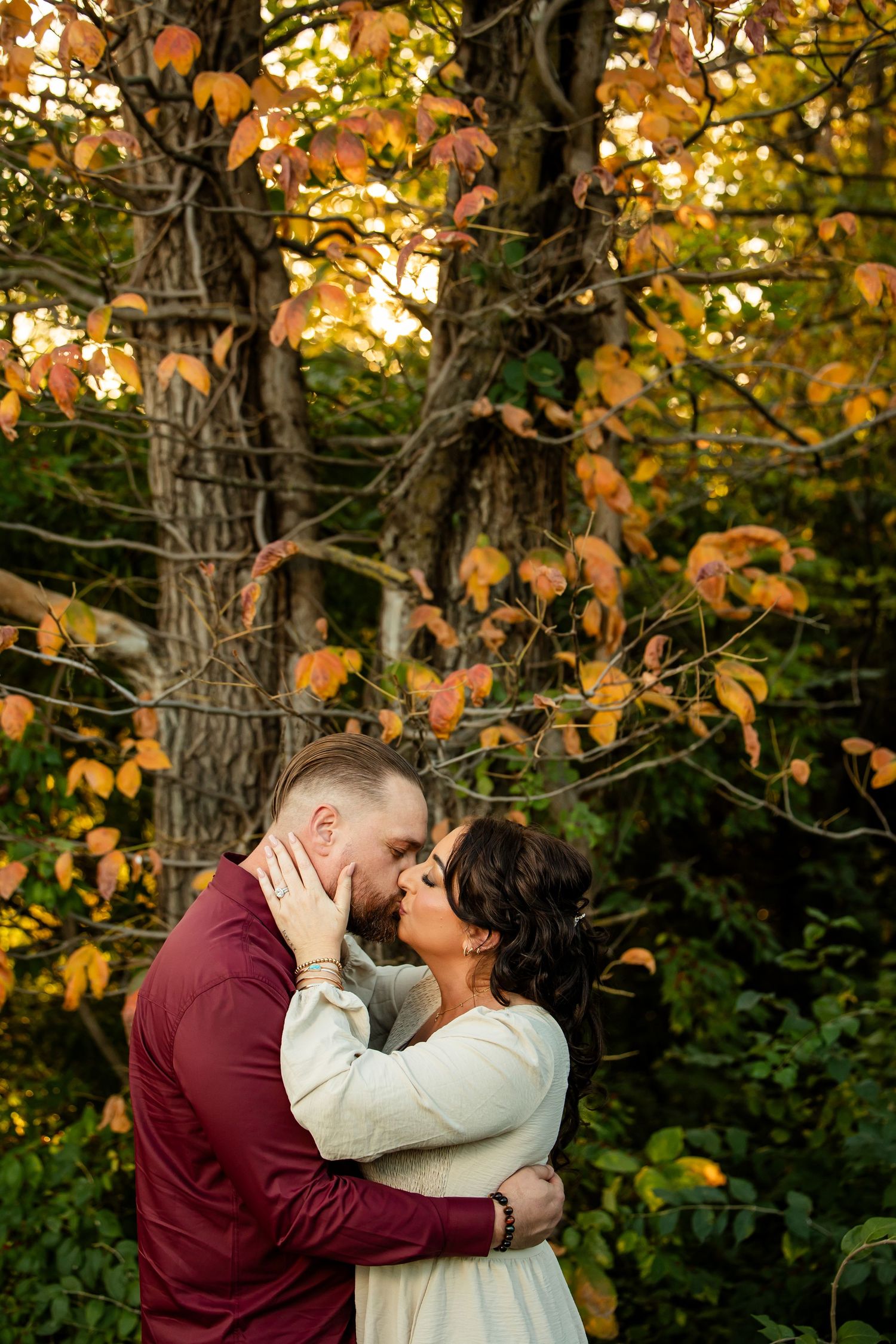Couple sharing a romantic kiss surrounded by vibrant autumn foliage and trees during a fall engagement photo session.