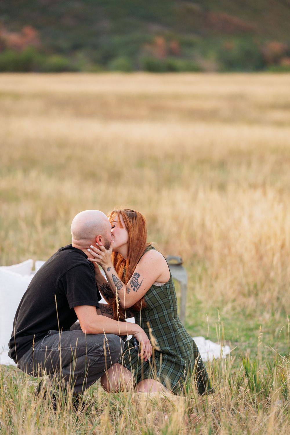 Couple sharing a romantic kiss while sitting on a blanket in a golden field with mountains in the background.