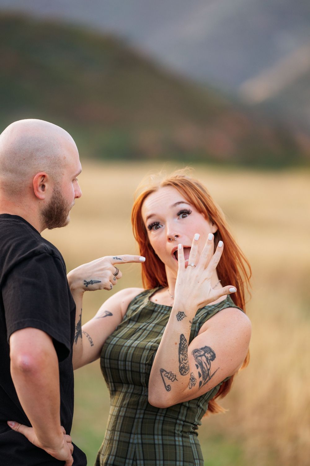 Tattooed redhead woman shows off engagement ring while bald bearded man points at it outdoors.