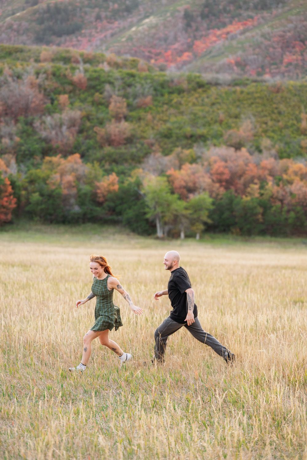 A couple runs hand-in-hand through a golden field with colorful autumn hills in the background.