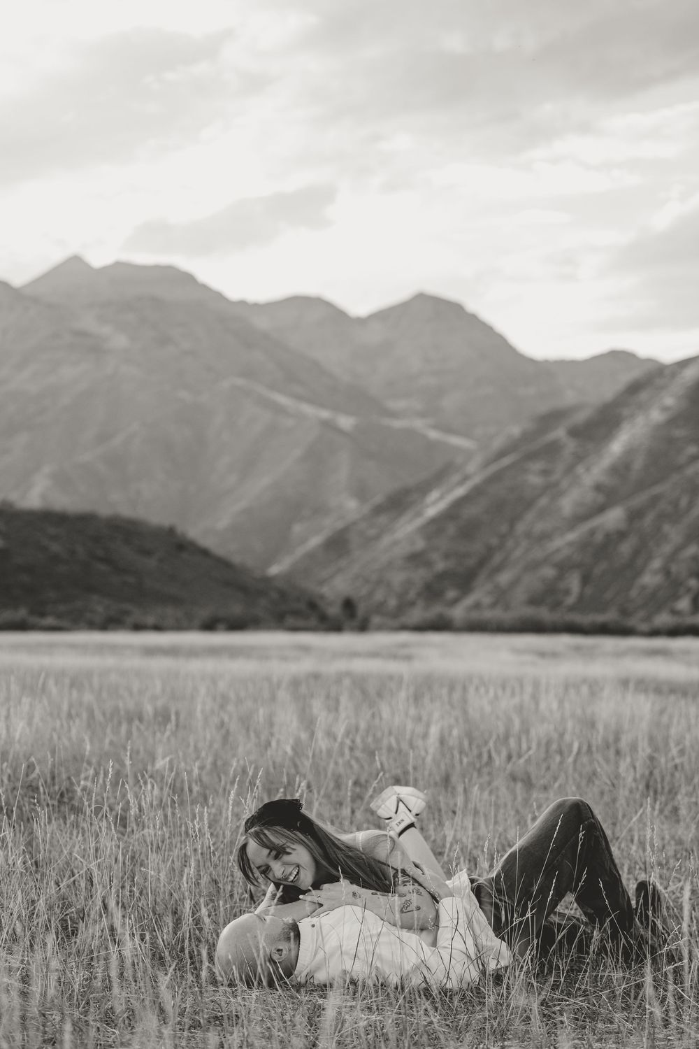 Black and white photo of a couple lying in a grassy field with mountains in the background.