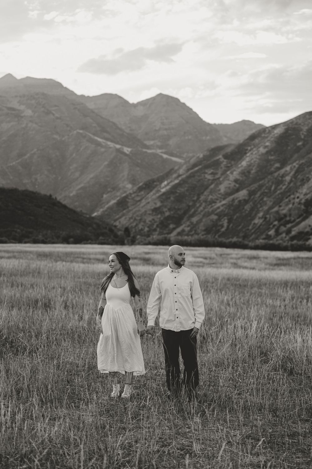 Black and white photo of a couple holding hands in a mountain meadow, woman in white dress looking back.