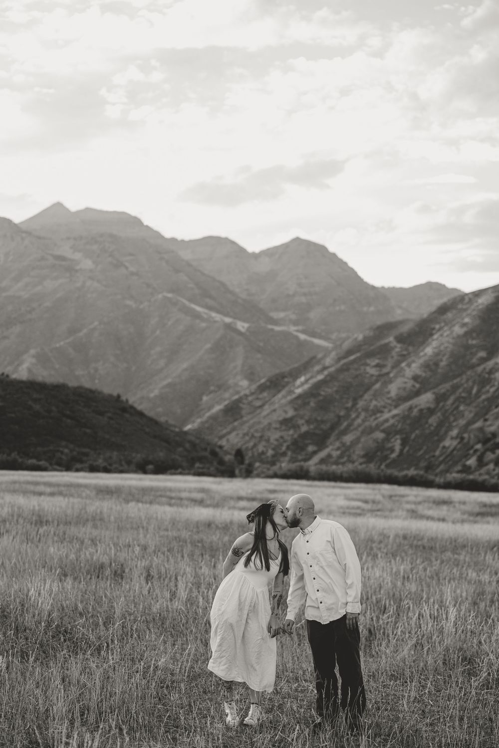 Black and white photo of a couple kissing in a mountain meadow, woman in flowing white dress, stunning peaks in background.