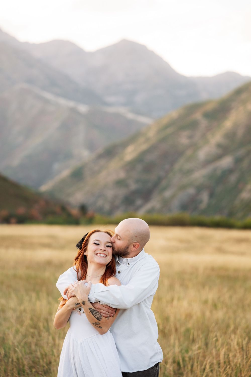 Couple embracing in golden field with mountain backdrop, man kissing woman's cheek, both wearing white.