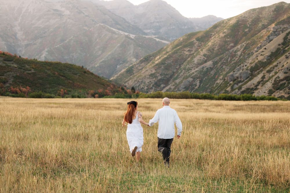 Couple holding hands walking through golden field with mountain backdrop in white outfits.