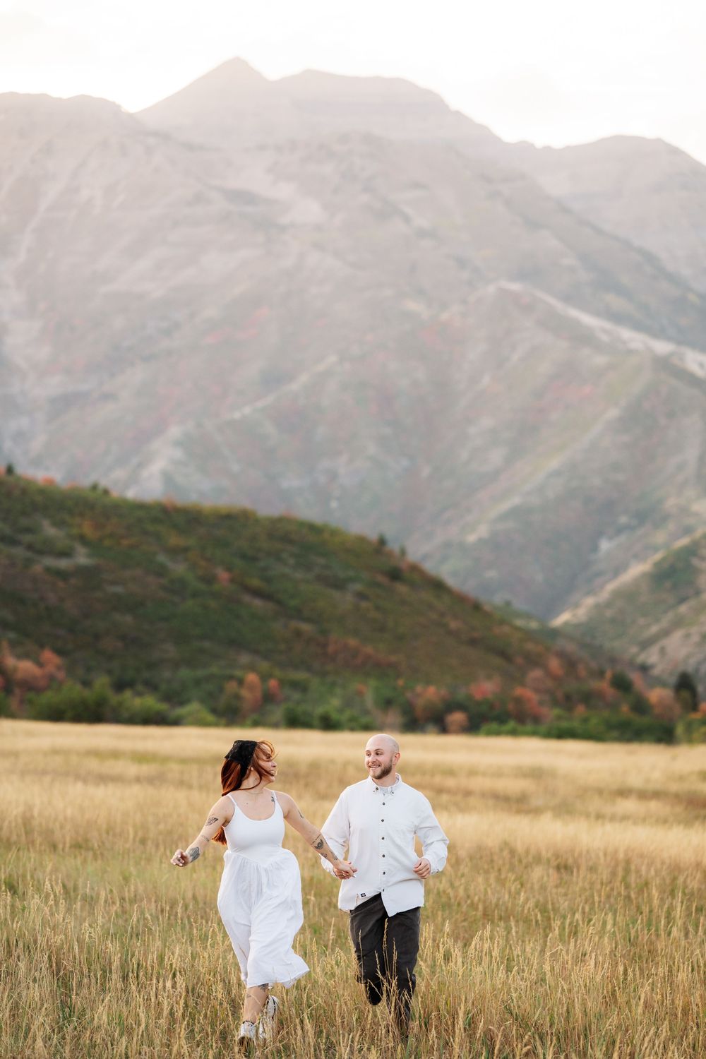 Couple holding hands running through golden field with stunning mountain backdrop in autumn.