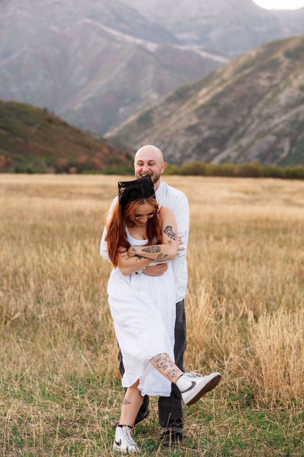 Tattooed couple embracing in golden field with mountain backdrop, woman in white dress lifted by bald man in white shirt.