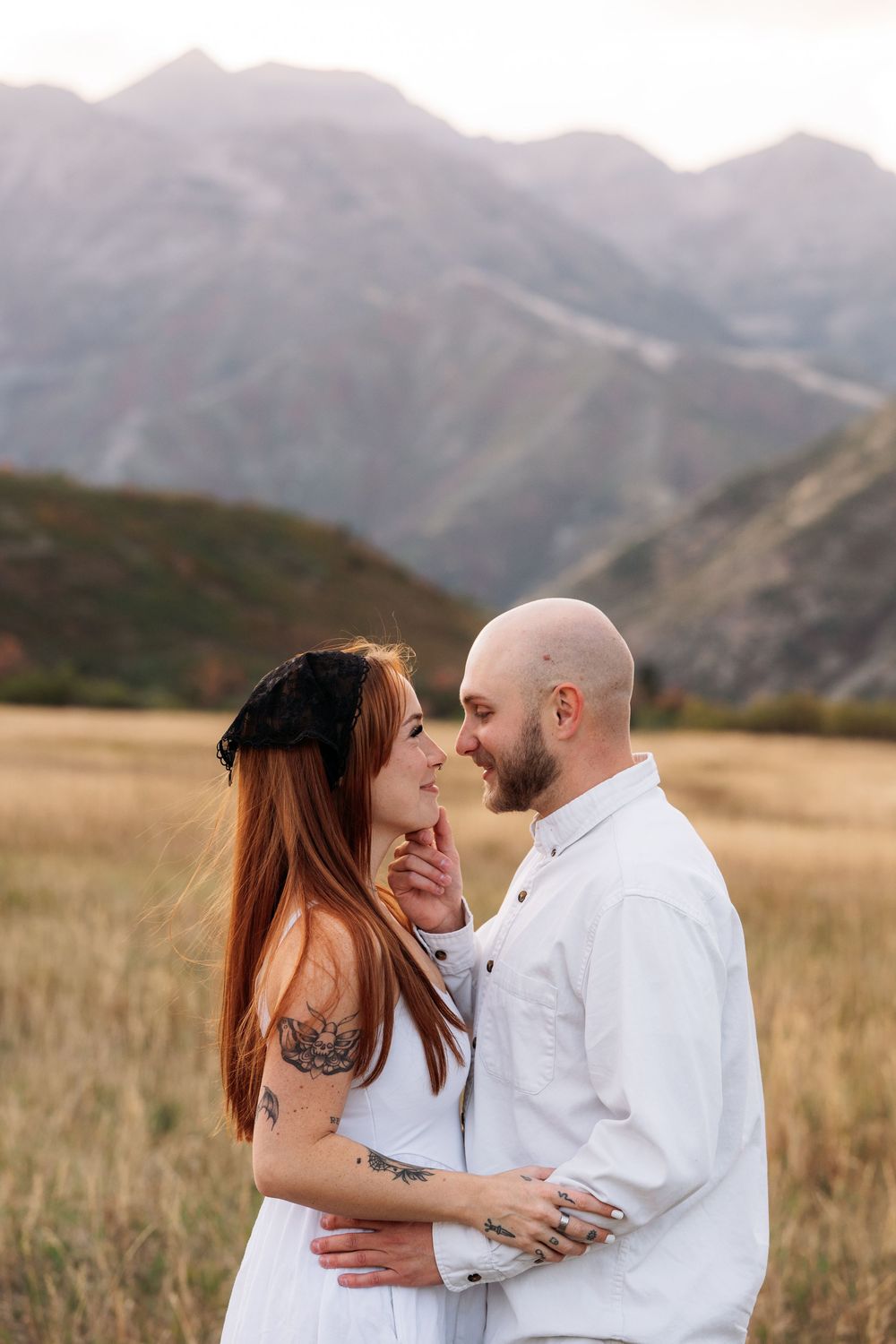 Couple embracing in golden field with mountain backdrop at sunset, woman with red hair and tattoos in white dress.