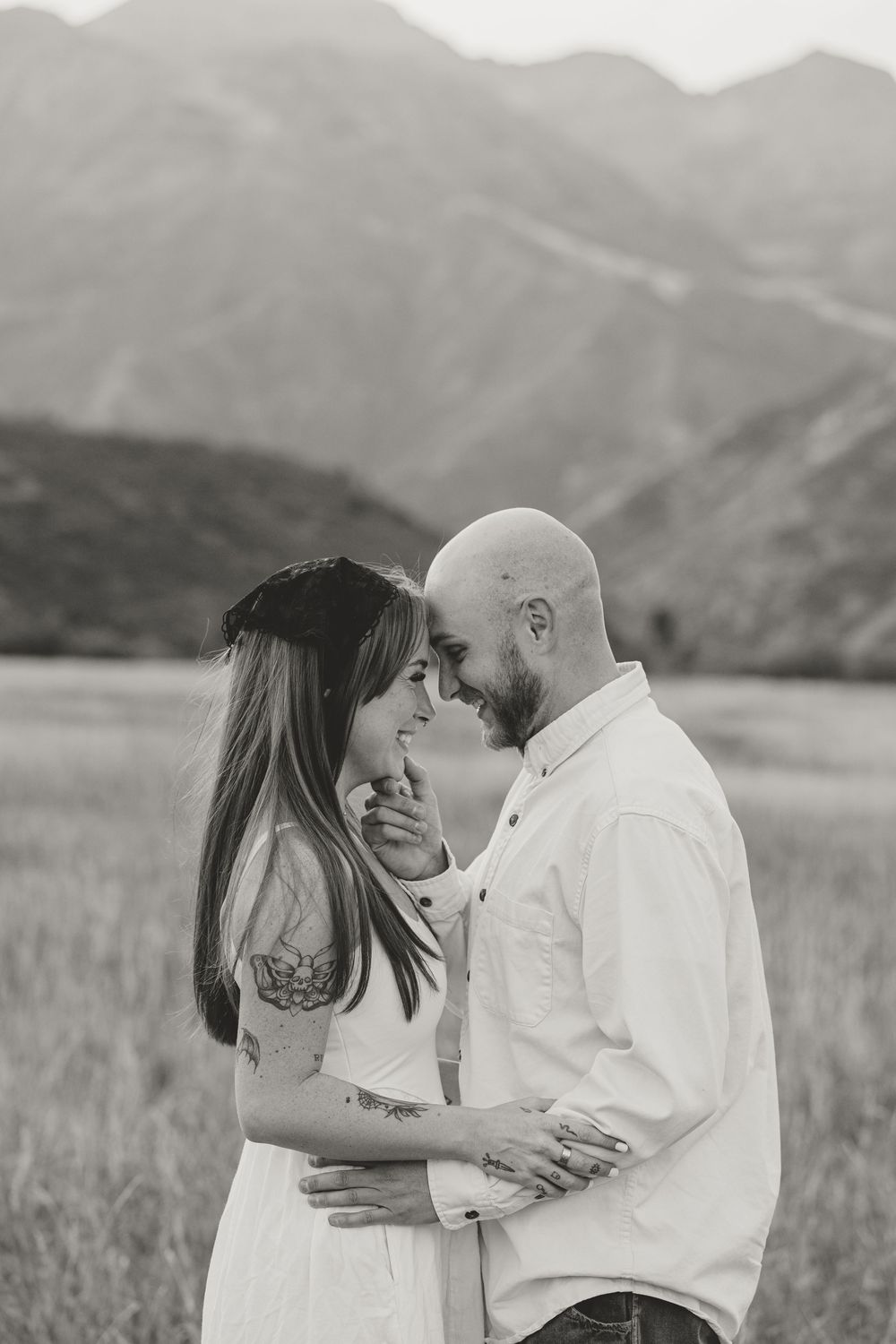 Black and white photo of a couple embracing in a field with mountains in the background.