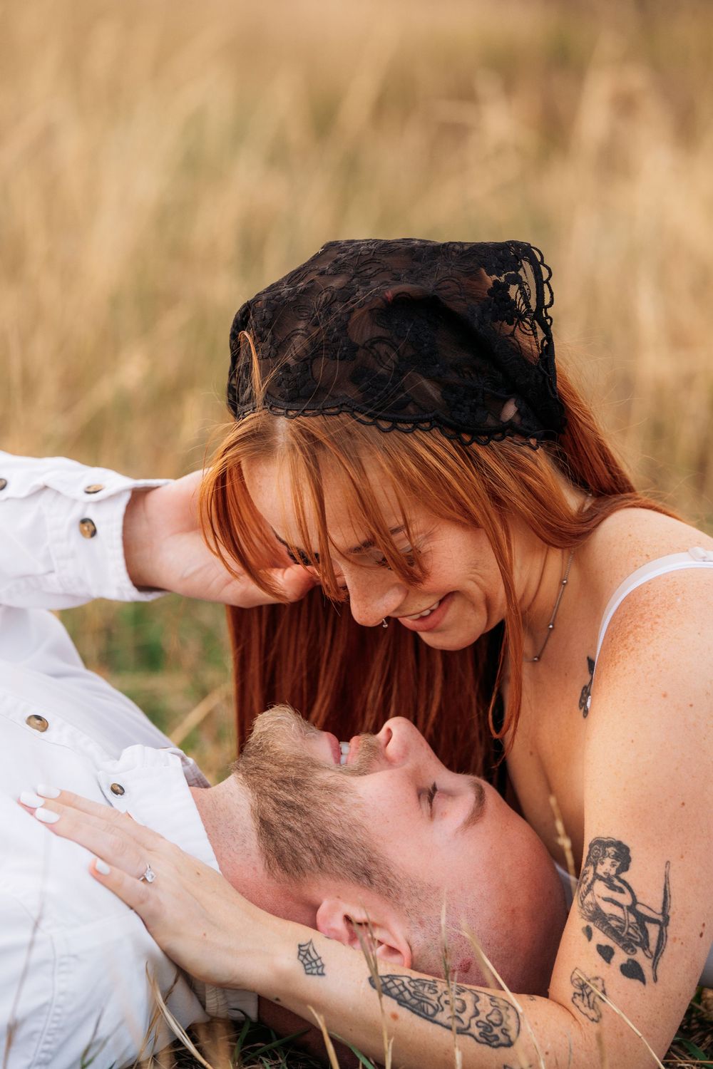 A redheaded woman with a black lace headpiece smiles at a tattooed man lying in a golden grass field.
