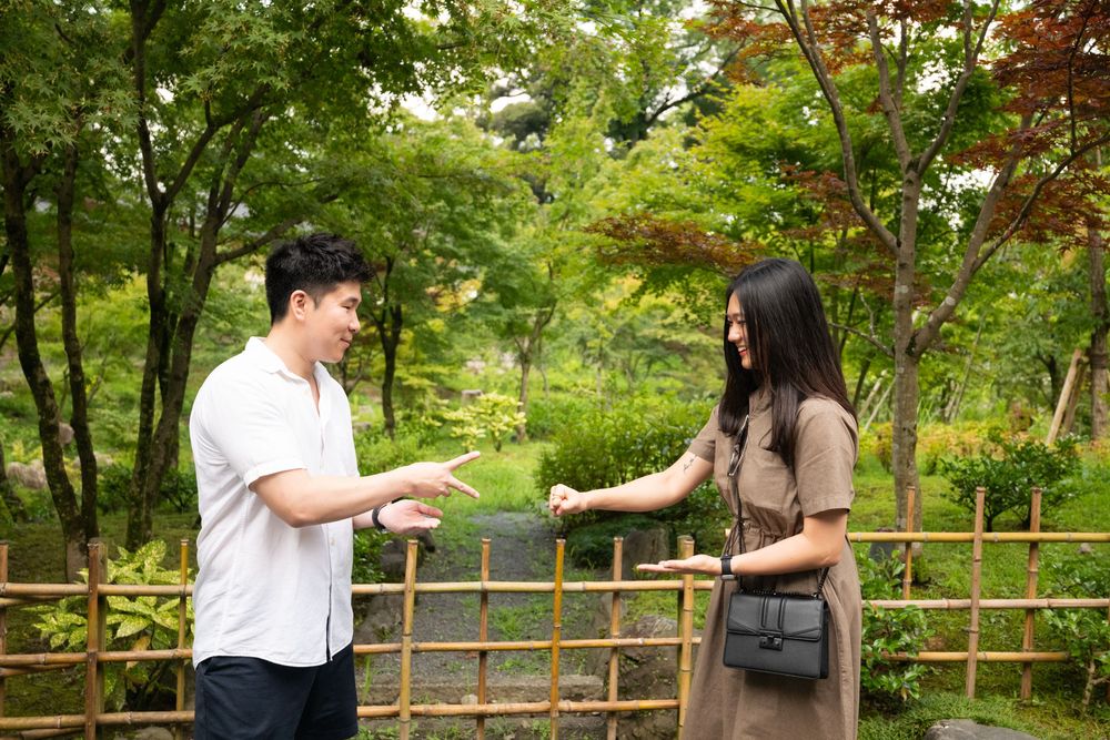 A smiling man and woman talk and gesture beside a wooden bamboo fence in a lush green Japanese garden surrounded by trees.