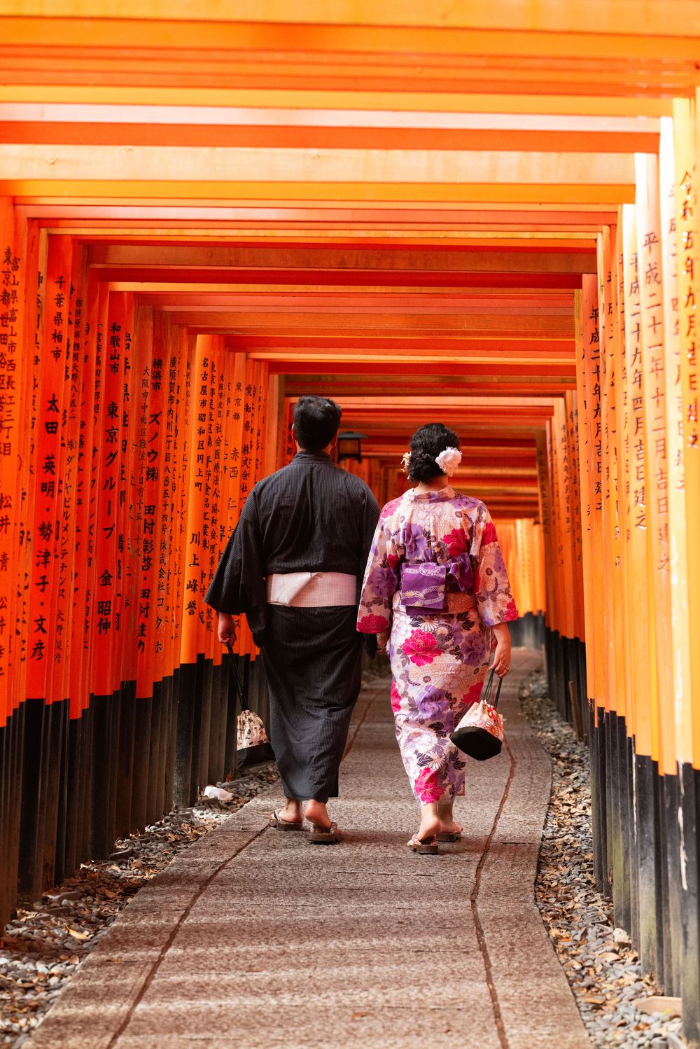 Couple in traditional kimono and yukata walking along a path beneath vermilion torii gates at Fushimi Inari Shrine in Kyoto, Japan.