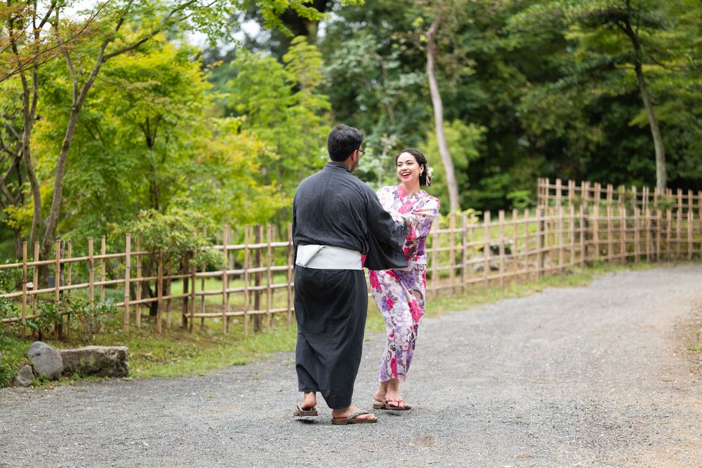 Couple in traditional Japanese yukata walking and smiling together on a gravel path beside a bamboo fence in a lush green garden.