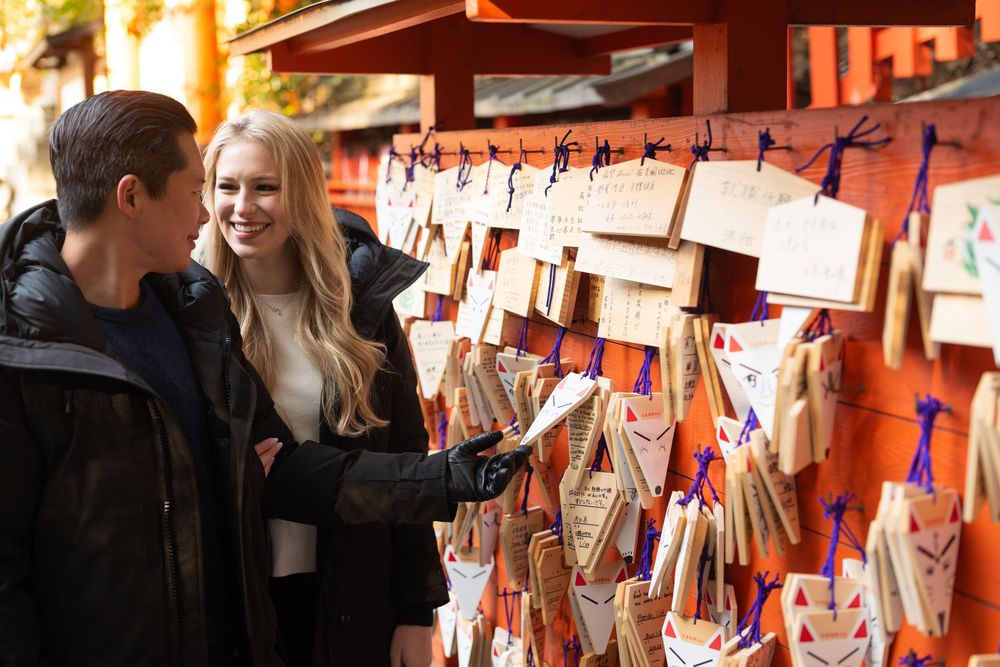 Smiling young couple in dark jackets browse wooden ema prayer plaques tied with ribbons at a Japanese shrine.