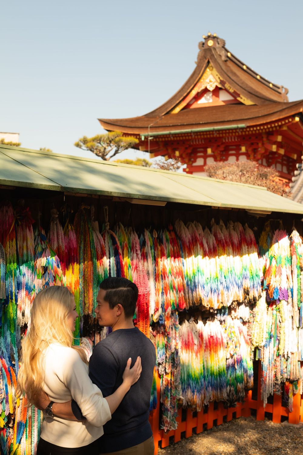 Couple embracing in front of colorful hanging origami paper cranes at a Japanese shrine with a red traditional temple roof behind them.