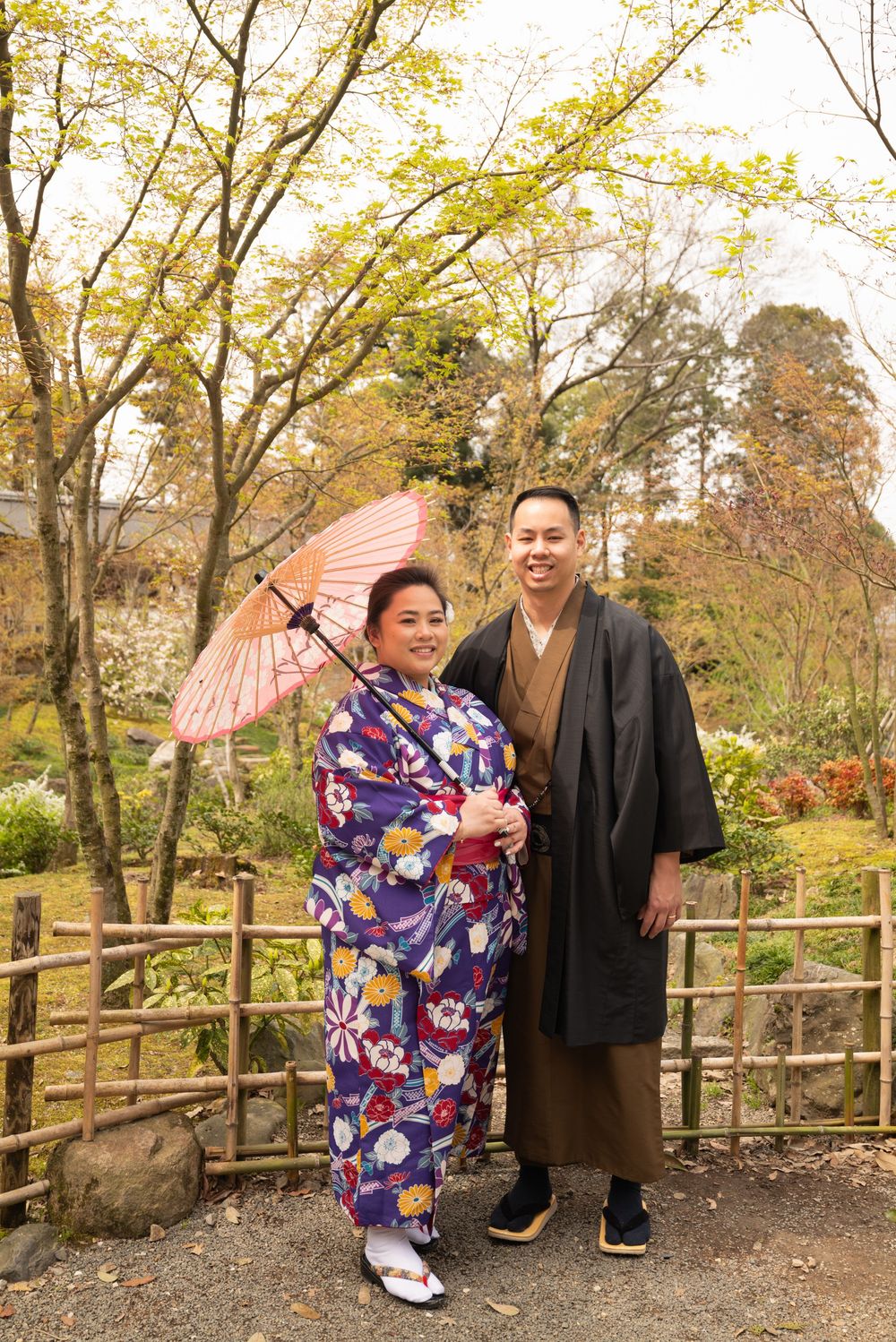 Couple in traditional Japanese kimono and haori posing in a spring garden, with a pink oil-paper parasol beside a bamboo fence.