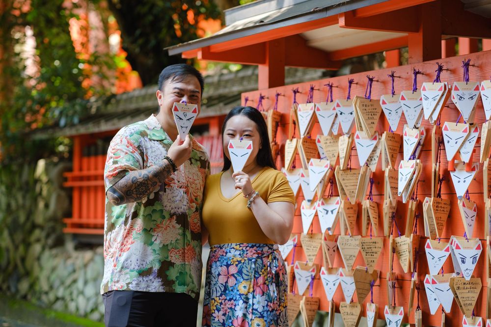 Smiling couple in colorful floral outfits hold small paper ema charms in front of a wall of hanging wishes at a Japanese shrine.