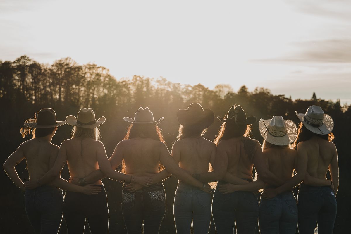 Group of women in cowboy hats stand arm in arm facing a glowing sunset over a treeline, seen from behind.