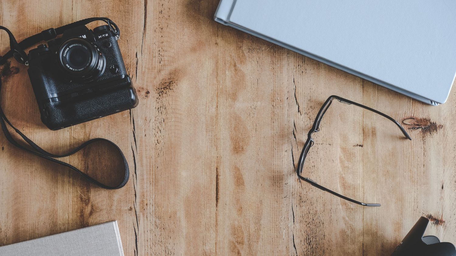 A vintage black camera with strap beside glasses and a laptop on a rustic wooden table surface.