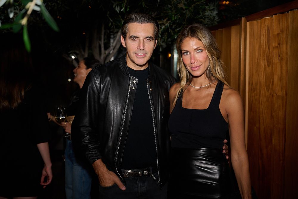 A man in a black leather jacket and a woman in a black sleeveless dress pose together at an evening party with guests in the background.
