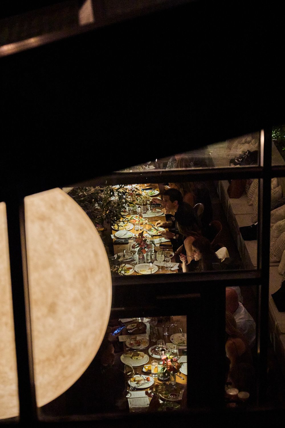 Diners gather around a long candlelit table set with plates and glasses, seen through a dark window in a cozy restaurant.