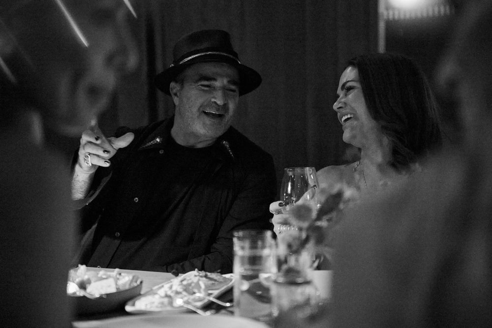Black and white photo of a smiling man in a hat and a laughing woman sharing drinks and food at a candlelit restaurant table.