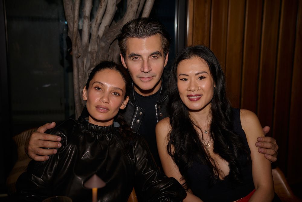 Three people pose closely together at a dimly lit restaurant, smiling at the camera in black outfits against a wood-paneled wall.